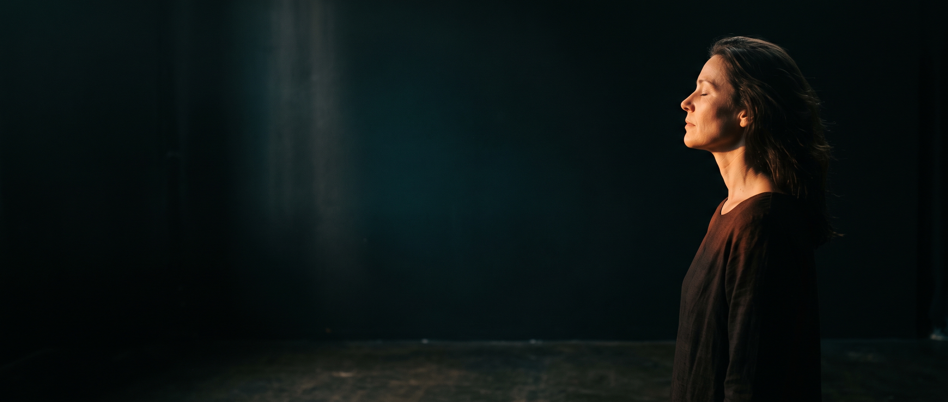 A woman in profile at the far edge of a dark studio, lit by one warm source